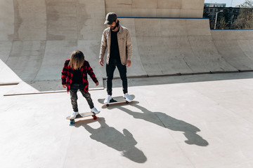 Young father and his son ride skateboards in a skate park with slides outside at the sunny day