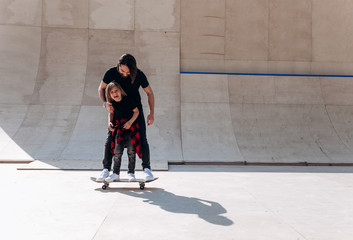 Father and his little son dressed in the casual clothes stand together on the one skateboard in a skate park at the sunny day