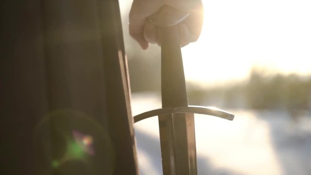 Man in medieval suit holding sword. Action. Close-up of man's hand in medieval suit holding sword hilt on background of sunlight. Epic view of medieval hero