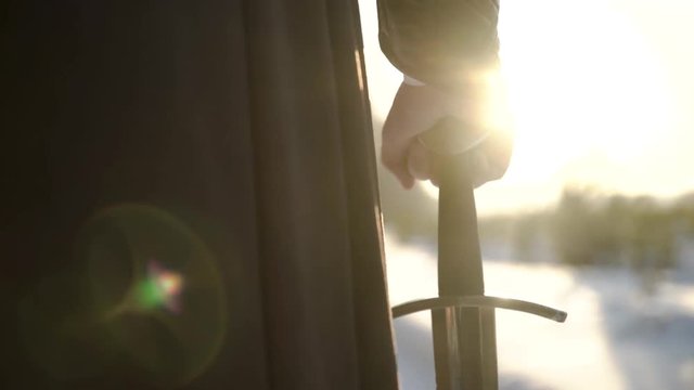 Man in medieval suit holding sword. Action. Close-up of man's hand in medieval suit holding sword hilt on background of sunlight. Epic view of medieval hero