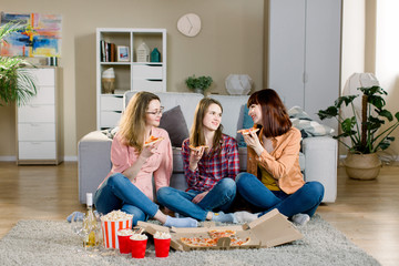 friendship, holidays, fast food and celebration concept - happy young women friends with drinks and popcorn eating pizza at home, sitting on the floor in cozy room