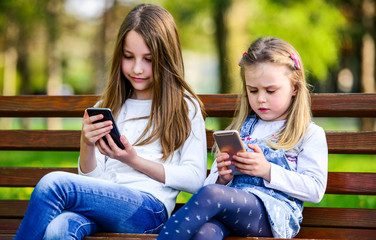 Two little girls having fun with smartphones in the park. Lifestyle, technology concept