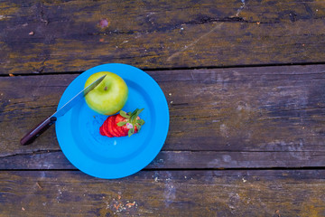 Fototapeta na wymiar green apple with red strawberries on a blue plate, with a brown wooden table background