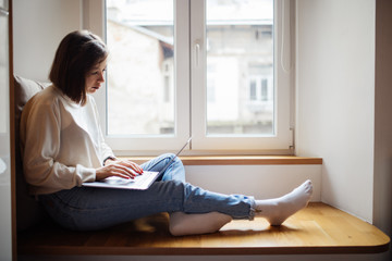 Beautiful young woman sitting on windowsill with laptop computer.