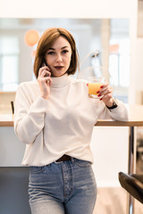 Smiling woman talking on mobile phone and holding glass of orange juice while having breakfast in a kitchen