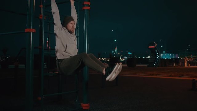 Bearded guy is hanging on training area in park in night and lifting legs