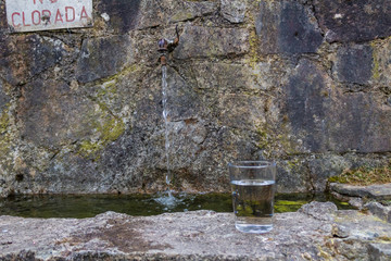 a full glass of water, leaning on a stone fountain, with a jet of water in the background
