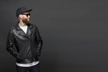 Man wearing leather biker jacket or asymmetric zip jacket with black cap, jeans and sunglasses. Handsome hipster man over gray background with copy space