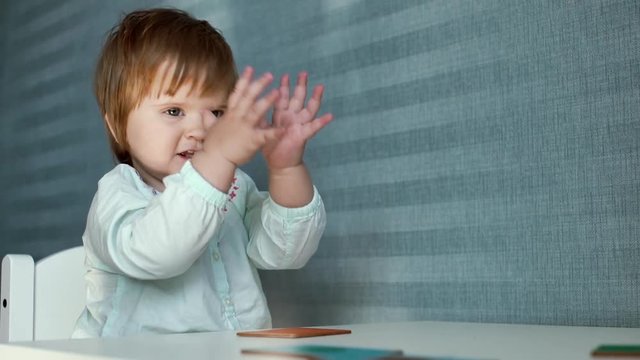 Little cute baby girl plays at small childs table and suddenly begins to clap her hands on the background of beautiful blue wallpaper in slow motion.