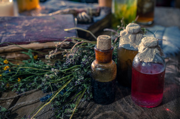 A witchery stuff: multiple tincture bottles, bunches of dry healthy herbs, stack of ancient ritual books, amulets, candles, chicken feet, sack of medicinal and magical herbs. 
