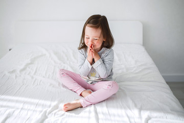 Little girl dreams with closed eyes. The child sits on a white bed and holds the lodoni by the face. The child practices yoga at home