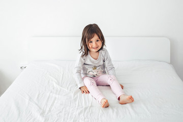 Naklejka premium Cheerful girl jumping on the bed. The girl laughs and jumps. On a white bed, the girl is having fun and jumping. Little baby in pink pants jumping on white bed 