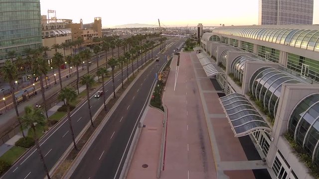 Aerial view along urban city center with few cars driving on mostly empty roads surrounded by palm trees, buildings, beautiful architecture, railroad tracks, and sidewalks at golden hour.