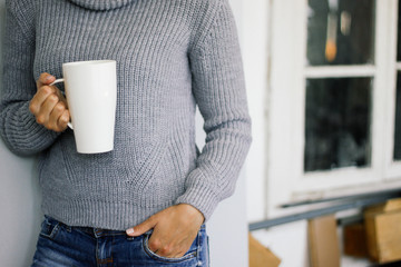 A woman in a warm gray sweater stands at the wooden window and holds a white mug in her hands. Style casual.