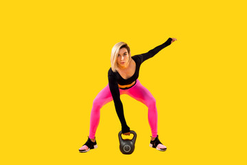 Fitness woman in fashionable pink and black sportswear work out with kettlebell on yellow background. Strength and motivation.