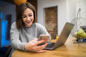 Young pretty woman sits at the kitchen table using a laptop and use on a cell phone.