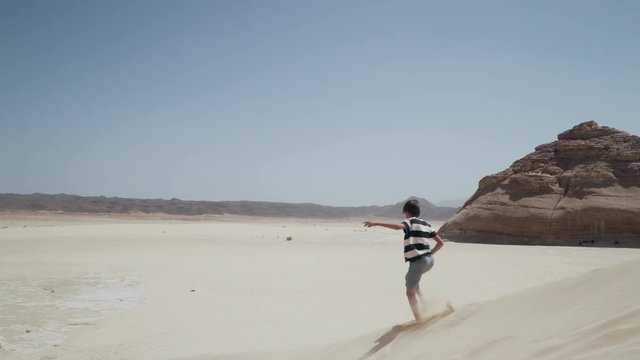 Attractive young boy running down by sand dune with rock in desert 4k