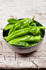 Fresh green raw peppers in metal bowl on rustic wooden table background.