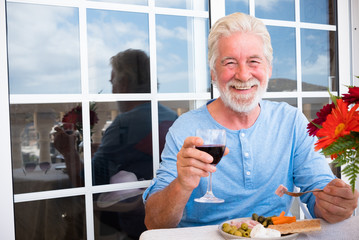 Happy senior man smiling enjoying a break with some food on the table. Sitting outdoor in terrace with a red wine glass. One caucasian people looking at camera with happiness