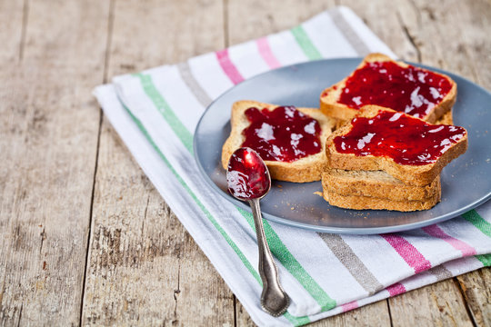 Fresh toasted cereal bread slices with homemade cherry jam and spoon on ceramic plate closeup on linen napkin on rustic wooden table background.