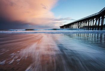 sunset on beach with pier during storm - obrazy, fototapety, plakaty