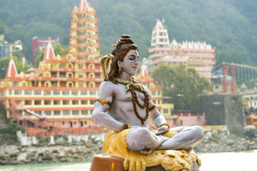 Stunning view of the statue of sitting Lord Shiva on the riverbank of the Ganges river. Blurred Trimbakeshwar Temple in the background. Rishikesh, Uttarakhand, India.