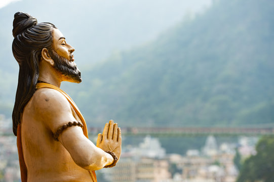 Stunning view of a sitting Hindu statue on the riverbank of the Ganges river. Blurred Lakshman Jhula bridge in the background. Rishikesh, Uttarakhand, India.