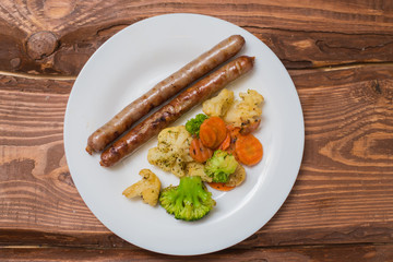Meat sausages and vegetables fried on a white plate on a wooden background.