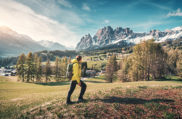 Obraz premium Young man in yellow jacket with backpack walking on the green meadow and looking on mountains at sunset in spring. Landscape with sporty guy, snowy rocks, trees, houses, blue sky in Italy. Travel