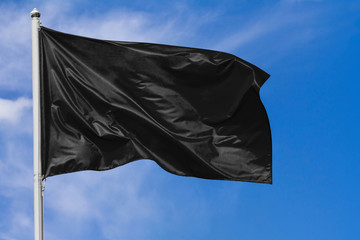 Black flag waving in the wind on flagpole against the sky with clouds on sunny day, closeup