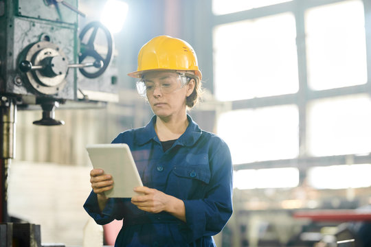 Waist up portrait of female worker wearing hardhat using tablet standing by machines in workshop, copy space