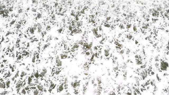 Top down aerial drone shot of big waves hitting the beach in bright sunlight leaving ever changing patterns on the water surface.