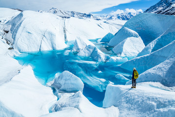 Young woman standing near deep blue lake on the Matanuska Glacier in Alaska. She wears a backpack and helmet with ice axe in hand for summer glacier travel. - obrazy, fototapety, plakaty