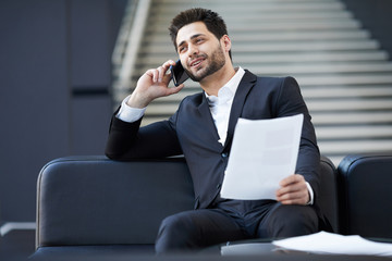 Content confident young mixed race businessman with beard sitting on leather sofa and discussing contract on mobile phone