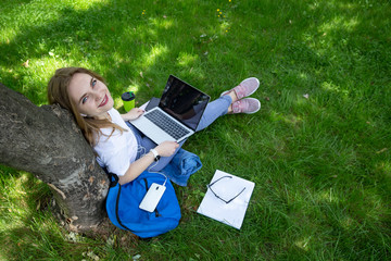 Freelance business concept. Top view of young woman who sits on the grass under tree with laptop on her legs. She look at camera and smile. Notebooks, coffee, dark-blue backpack, phone near. - obrazy, fototapety, plakaty