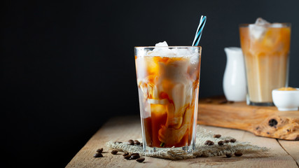 Ice coffee in a tall glass with cream poured over, ice cubes and beans on a old rustic wooden table. Cold summer drink with tubes on a black background with copy space