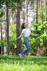Naklejka premium Happy laughing blonde girl with white t-shirt, blue jeans and rose sneakers is jumping up on the green grass at the summer park.