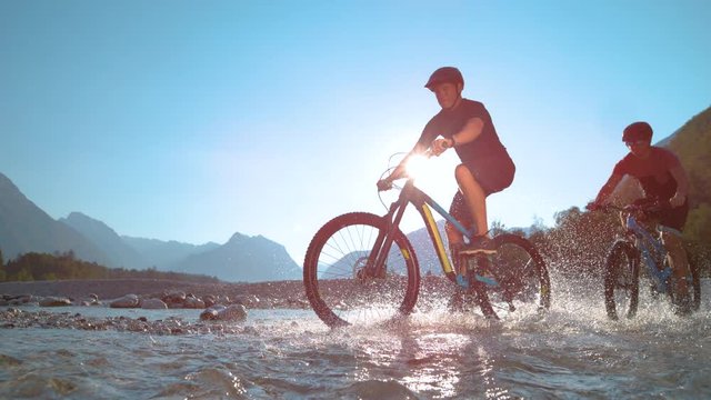 SUPER SLOW MOTION, LOW ANGLE: Guys riding ebikes along the shallow river and splashing refreshing water on a sunny day in beautiful Slovenian nature. Young friends spraying water during a bike ride.
