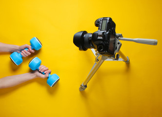 Fitness blogger. Woman Holding plastic dumbbells with her hands and blogging with camera on tripod on yellow background. Top view