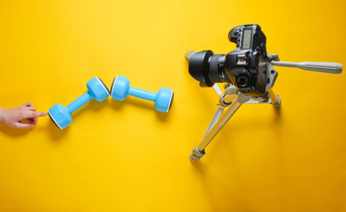 Fitness blogger. Woman Holding plastic dumbbells with her hands and blogging with camera on tripod on yellow background. Top view