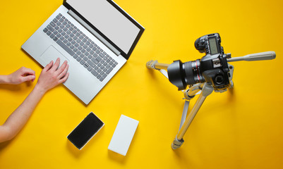 Tehnobloger concept. Women's hands unboxing new smartphone with box and blogging with camera on tripod, laptop on yellow background. Top view
