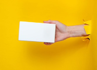 Female hand holds white box through torn yellow paper. Minimalistic creative fashion concept