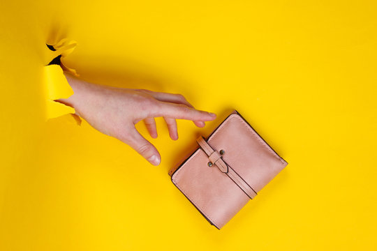 Female hand holding pink wallet through torn yellow paper. Minimalistic creative fashion concept