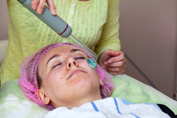 A young girl is lying on couch with a pink hat on her hair while performing an darsonvalization on face with a special device while cleaning and removing body fat under the skin on nose. Cosmetology