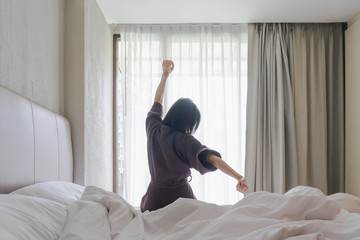 Woman stretching in bed after waking up, back view. Woman sitting near the big white window while stretching on bed after waking up with sunrise at morning, back view.