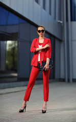 high fashion outdoor portrait of a young woman in a red pantsuit. Stylish black sunglasses, a black clutch bag, long hair.