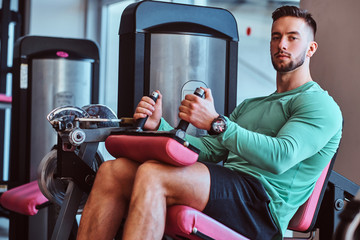 Strong pensive man is sitting on training apparatus in gym and doing legs exercises.