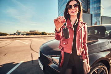 Happy smiling woman holding key for car, which is behide her, she just made a good deal seiling her car.
