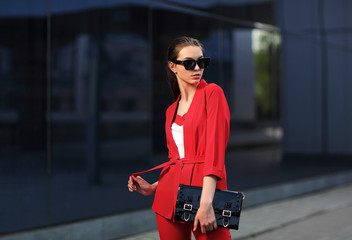 high fashion outdoor portrait of a young woman in a red pantsuit. Stylish black sunglasses, a black clutch bag, long hair.