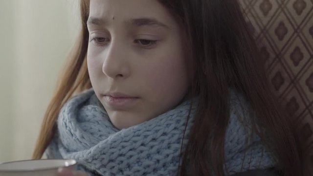 Portrait of teenage girl wrapped in warm scarf holding a cup of hot tea in hands. The girl feels bad, she is ill and has a fever. Concept of healthcare, treatment